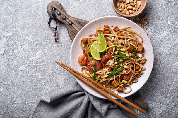 Top view of plate with asian udon noodles, spicy shrimps, fried tofu cheese, peanuts, lime and green onion on grey concrete background - Photo, image