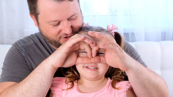 Mała córeczka 3 lata i ojciec bawią się w domu, robiąc śmieszne miny, robiąc okulary palcami, jak okulary patrzące w kamerę przez lornetkę. baw się z dzieckiem, za darmo - Zdjęcie, obraz