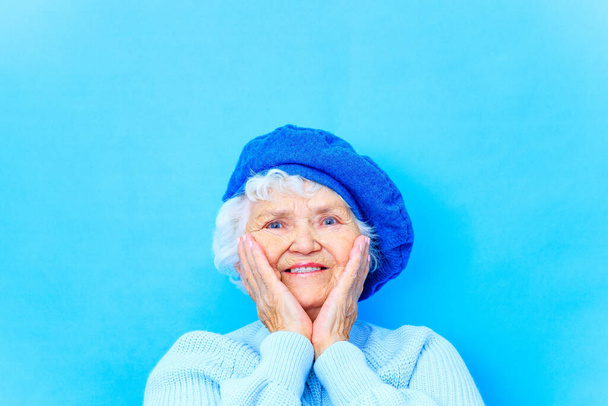 schöne Rentnerin in blauer Baskenmütze und warmem Pullover blickt in die Kamera und lächelt im blauen Wandhintergrund im Studio - Foto, Bild