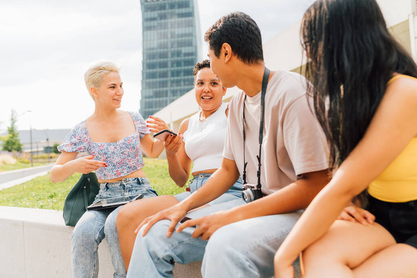 Grupa młodych wielonarodowych przyjaciół siedzi na świeżym powietrzu i rozmawia ze sobą, dobrze się bawiąc, spędzając razem czas - Zdjęcie, obraz