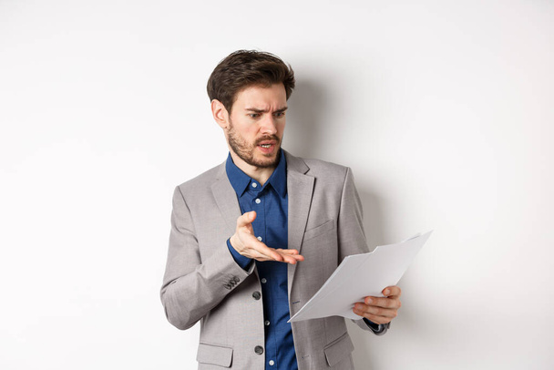Naštvaný zaměstnavatel při pohledu na špatný dokument, stěžuje si na dohodu, ukazuje na papír frustrovaný, stojí v obleku na bílém pozadí - Fotografie, Obrázek