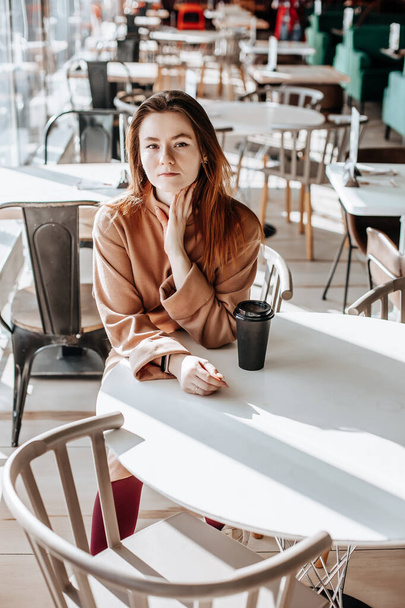 Stylish girl sits in a cafe and drinks coffee. Coffee to go in a cardboard cup. Woman with ginger hair in a beige warm suit in a cozy atmosphere. Modern interior. Calm and pleasant pastime - Photo, Image