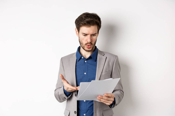 Homme d'affaires confus regardant un mauvais document, se plaignant sur l'affaire, pointant du doigt le papier frustré, se tenant en costume sur fond blanc - Photo, image