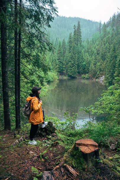 woman hiker with backpack in yellow raincoat looking at mountain lake copy space - Fotografie, Obrázek