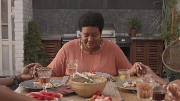 Mellkas-up érett afro-amerikai nő ül az asztalnál, kéz a kézben együtt levágott családtagok, imádkozva vacsora előtt - Felvétel, videó