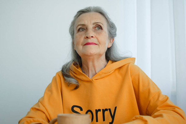 Close-up portrait of pensive and calm mature woman with gray hair and holding a coffee mug - Photo, Image