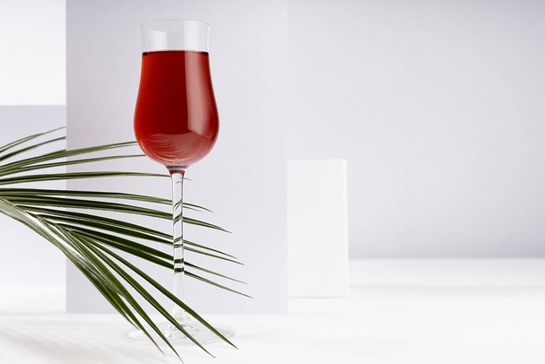 Red liquor in rich glass in sunlight with shadows, green palm leaves as decoration in soft light white abstract interior with geometric spaces. - Fotoğraf, Görsel