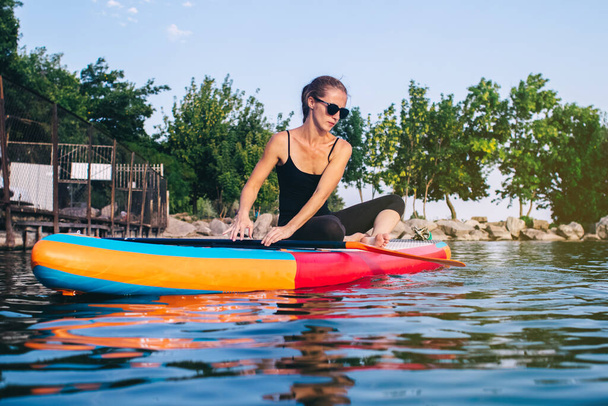 Close-up of a girl doing yoga on sub board. Psychology meditation, relaxation and self-healing concept. Lonely woman alone on board against a background of water and a pier. Stand up paddle boarding - Photo, Image
