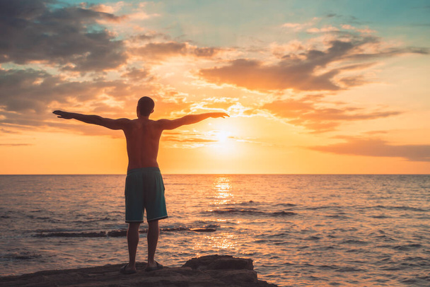 Silhouette of a young man raising his arms wide open on the sea side of relaxing day at dawn surrounding by nature. Meditation and concentration posture. Freedom and relax vacations concept.  - Foto, immagini