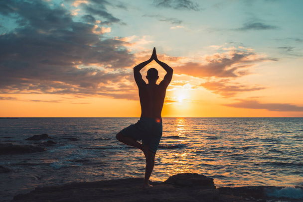 A fit young man practices sun salutation yoga on the beach during sunset. Meditation and concentration posture on the sea side of relaxing day at dawn surrounding by nature. - Valokuva, kuva