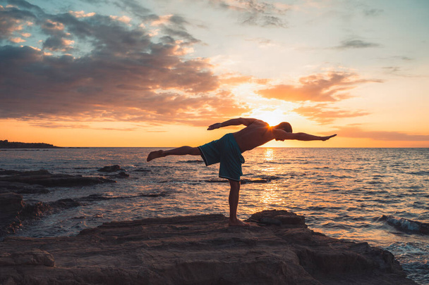 A fit young man practices yoga poses on the beach during sunset. Meditation and concentration posture on the sea side of relaxing day at dawn surrounding by nature. - Фото, изображение