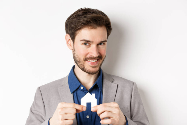 Pojetí nemovitostí a pojištění. Usmívající se muž realitní makléř ukazující malý papírový dům výřez a při pohledu do kamery, prodej nemovitostí, bílé pozadí - Fotografie, Obrázek