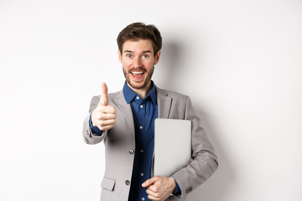 Good job. Happy smiling business man in suit holding laptop, showing thumb up to praise you, nice work gesture, standing on white background - Foto, Imagem