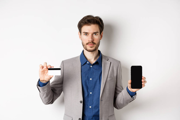 Интернет-магазины. Серьёзный бородатый мужчина в деловом костюме показывает пластиковую кредитку с пустым экраном смартфона, стоящую на белом фоне - Фото, изображение