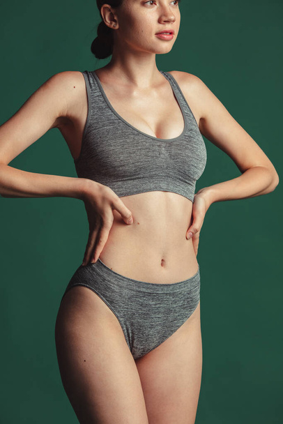 Vista de perfil de cuerpo delgado perfecto. Mujer joven con su piel sana natural, usando ropa interior de algodón gris sobre fondo verde - Foto, Imagen