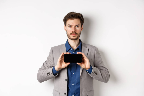 E-commerce et concept d'achat en ligne. Jeune homme sérieux en costume montrant écran vide smartphone horizontal, debout sur fond blanc - Photo, image