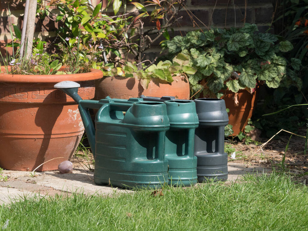 Três latas de rega verdes estão lado a lado em lajes prontas para regar um jardim. Vasos de flores atrás e grama em primeiro plano. O bico de rosa pode ser visto em uma lata. - Foto, Imagem