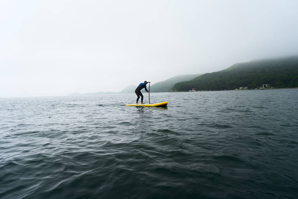Man in blue wetsuit is paddling on a yellow SUP board on a sea waves at cloudy day. - Foto, Imagen