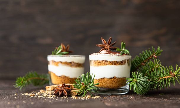 Layered dessert in glass jar with cookie crumble and whipped cream decorated with rosemary and anise, dark background. No bake cheesecake, trifle or pudding. - Fotoğraf, Görsel