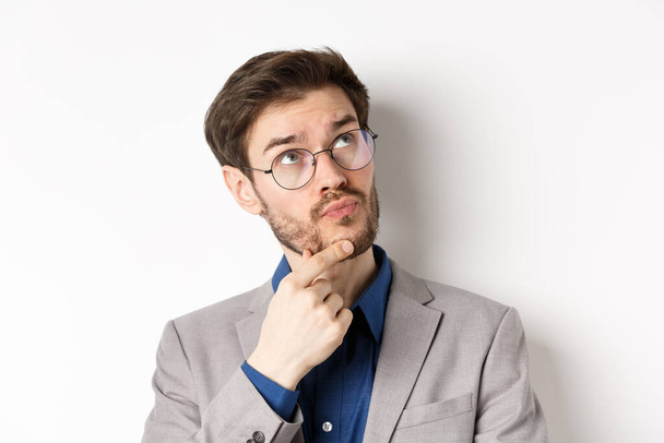 Thoughtful businessman with beard and glasses making decision, looking up and thinking, standing in suit with pensive face, white background - Foto, Imagem