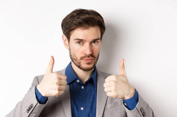 Well done. Satisfied businessman in suit showing thumbs up in approval, like and agree, recommend good company, white background - Foto, imagen