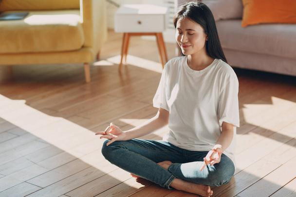 Femme asiatique faisant du yoga et zen comme la méditation pose de lotus en tenue décontractée à l'intérieur salon appartement avec lumière naturelle du soleil - Photo, image