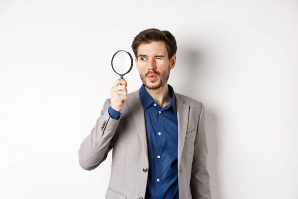 Handsome male model in suit looking through magnifying glass with interest, seeing something aside, white background - Foto, Imagem