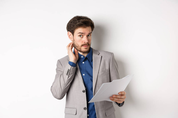 Confused office worker in suit scratching beard and look clueless, cant understand document, holding paper with indecisive face, white background - Photo, Image