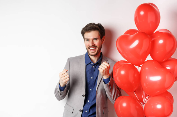 Lucky handsome man dancing and saying yes, standing near lovers day heart balloons and smiling, standing against white background - Photo, Image