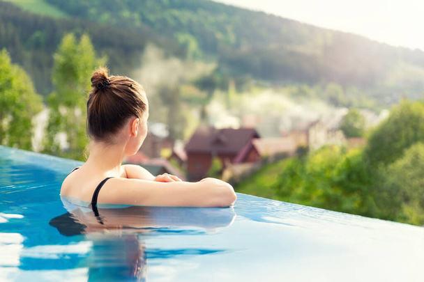 back view young adult female person enjoy relax in infinity edge luxury outdoor swimming pool looking on fog hill green mountains in warm sunset light. Alpine welness travel vacation tourism concept - Photo, Image