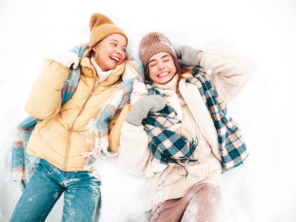 Two young beautiful smiling hipster female in trendy warm clothes and scarfs.Carefree women posing in the street in park. Positive pure models lying in snow. Enjoying winter moments. Xmas concept - Φωτογραφία, εικόνα