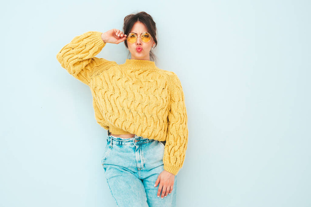 Young beautiful smiling female in trendy summer yellow hipster sweater and  jeans. Sexy carefree woman posing near light blue wall in studio. Positive model having fun indoors. Makes duck face - 写真・画像