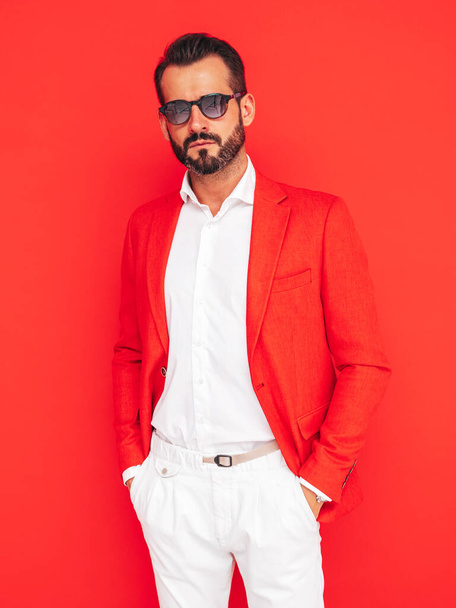 Portrait of handsome confident stylish hipster lambersexual model. Sexy modern man dressed in elegant suit. Fashion male posing in studio near red wall in sunglasses - Фото, изображение