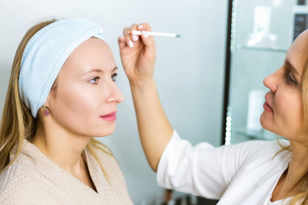 Hautpflege und Facelifting. Eine junge Frau bei einem Kosmetiktermin. Ein Fachmann untersucht es und macht mit einem speziellen Bleistift Markierungen - Foto, Bild