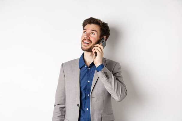 Unbekümmerter junger Mann im Handy-Gespräch und lächelnd, mit verträumtem Gesicht aufblickend, im Anzug auf weißem Hintergrund stehend - Foto, Bild