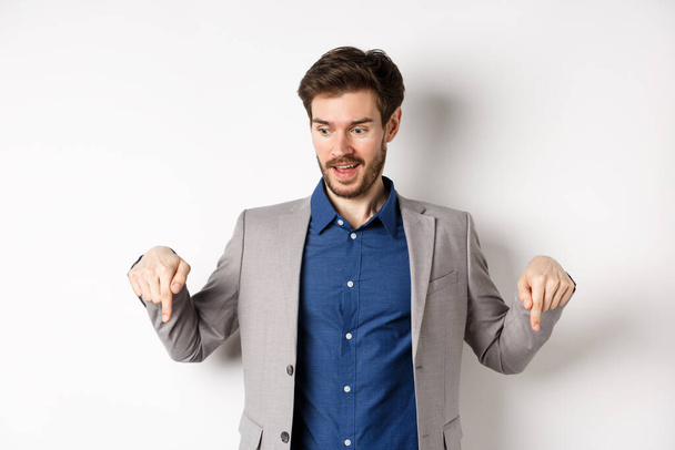 Opgewonden man in pak glimlachend en wijzende vingers naar beneden, op zoek naar speciale deal met geamuseerd gezicht, staande op witte achtergrond - Foto, afbeelding
