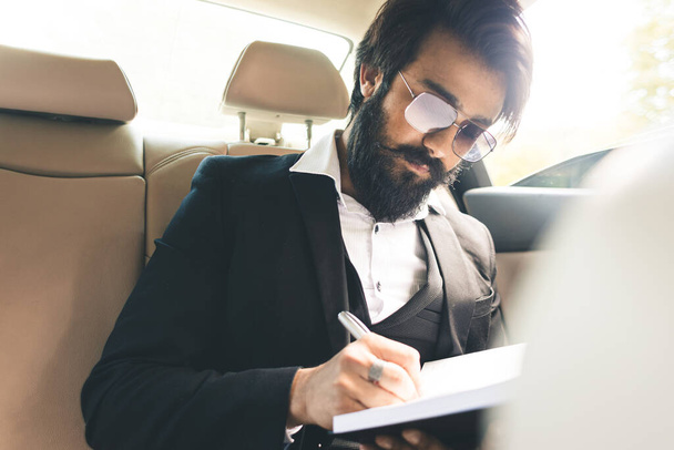 A stylish and successful Indian with glasses and a beard works and plans his schedule in the back seat of the car. Safe and comfortable business travel - Fotoğraf, Görsel
