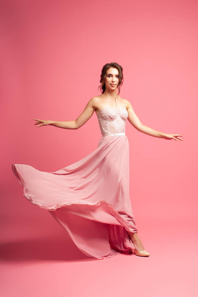 full-length portrait of smiling dark-haired woman in long pink evening dress, with skirt flying in motion, isolated on pink background - Photo, image