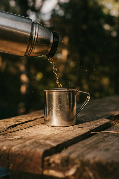 Tea is poured into a metal mug from a traveling metal thermos. Rest of the tourist in the forest. A tourist pours hot tea from a thermos into a mug. Tourist breakfast - Fotó, kép