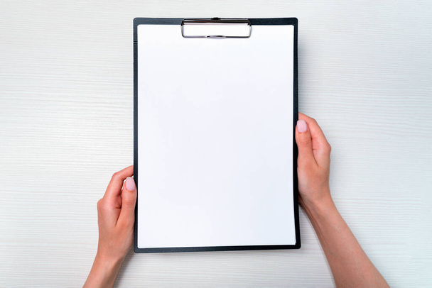 mockup female hands hold clipboard with a4 paper over light wooden table. top view close-up. flat lay. - 写真・画像