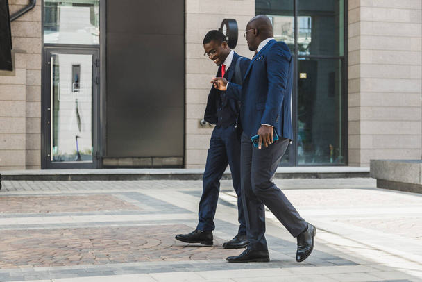 Dwóch ciemnoskórych afrykańskich biznesmenów w garniturach i okularach spaceruje po mieście i rozmawia o interesach. - Zdjęcie, obraz