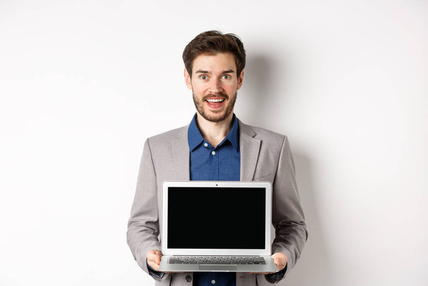Knappe blanke zakenman in pak met lege laptop scherm, promo demonstreren, staan op witte achtergrond - Foto, afbeelding
