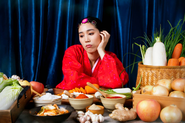 Hermosa mujer asiática con un hanbok rojo está sentada, considerando y encontrando métodos de cocina de verduras frescas para preparar recetas de comida tradicional coreana para Kimchi en su casa: enfoque selectivo - Foto, Imagen