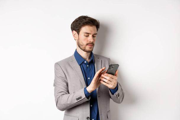 Mladý podnikatel v obleku textové zprávy na mobilním telefonu, při pohledu na smartphone, stojí na bílém pozadí - Fotografie, Obrázek