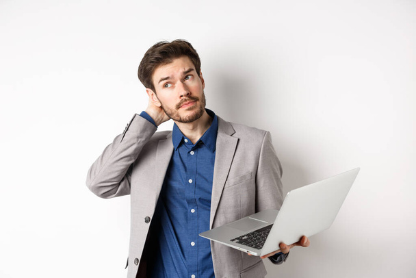 Задумчивый бизнесмен царапает голову и отворачивается, принимая решение во время работы на ноутбуке, стоя на белом фоне - Фото, изображение