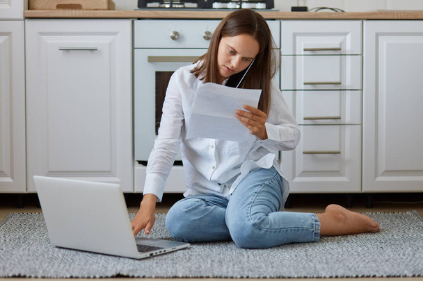 Indoor shot of concentrated female wearing white shirt and jeans, holding paper sheet and talking phone, working on computer, posing on floor in kitchen. - Photo, image