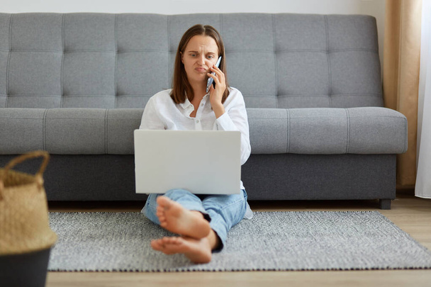 Portrait of young adult female wearing white shirt and jeans sitting on floor near sofa, working on laptop computer and talking phone with upset facial expression. - Фото, изображение