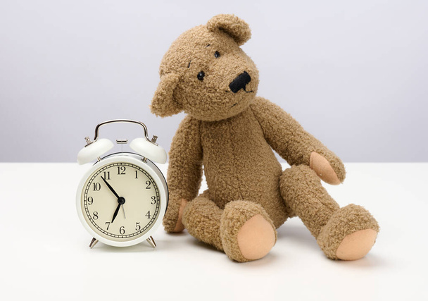 Brauner Teddybär sitzt auf einem weißen Tisch und einem runden Wecker, morgens um fünf vor sieben. - Foto, Bild
