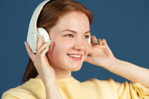 Close-up portrait of young red-haired girl in headphones looking away isolated over blue background. - Φωτογραφία, εικόνα