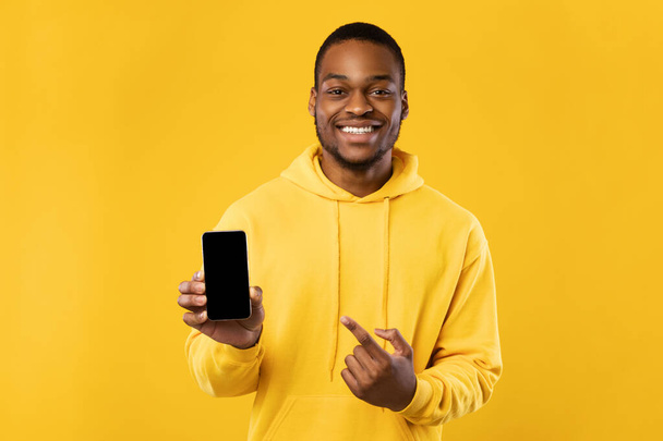Schwarzer Mann zeigt Handy mit Zeigefinger auf Bildschirm, gelber Hintergrund - Foto, Bild